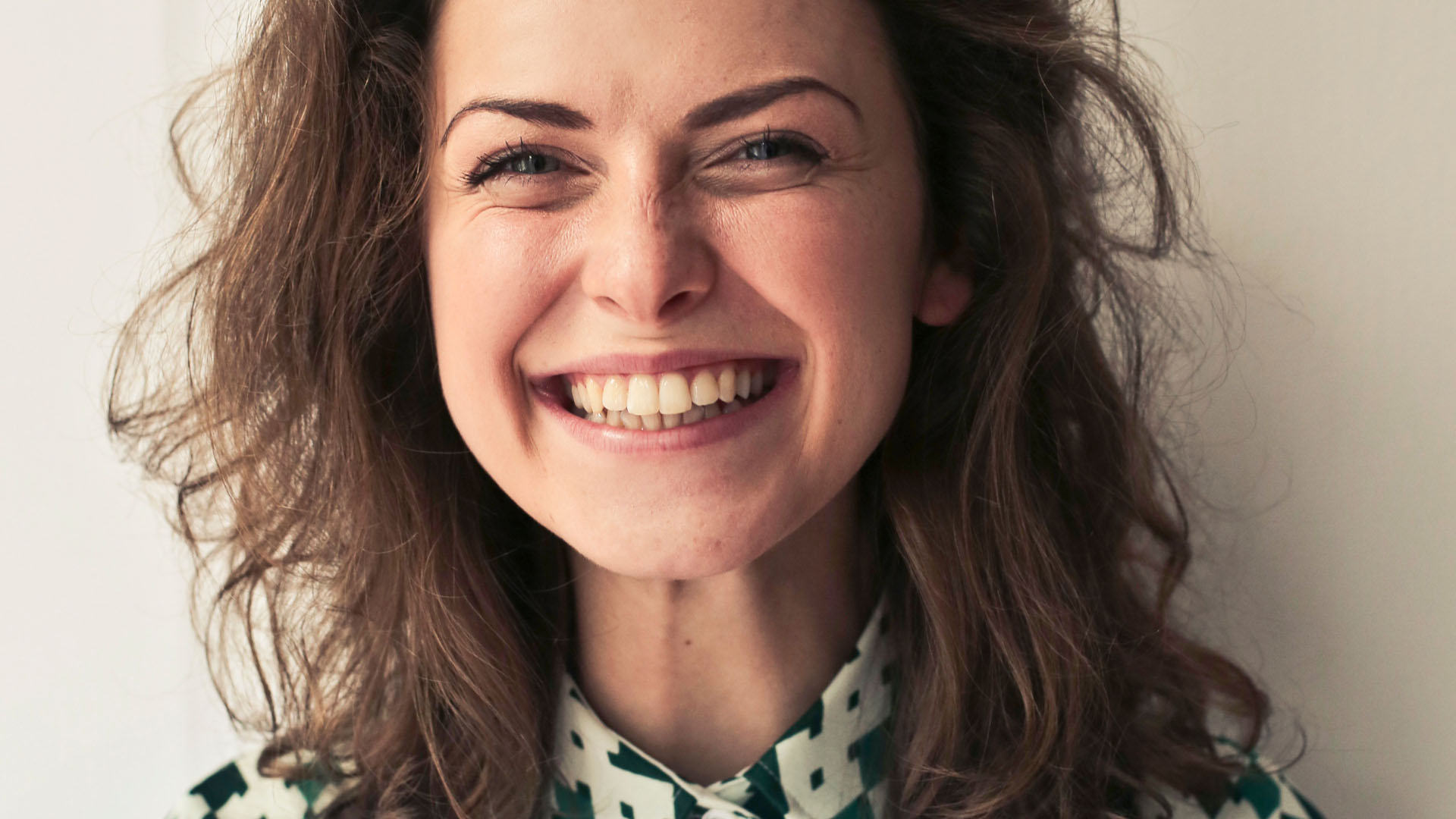 Young woman with curly brown hair smiling broadly, wearing a green and white patterned shirt against a light background.